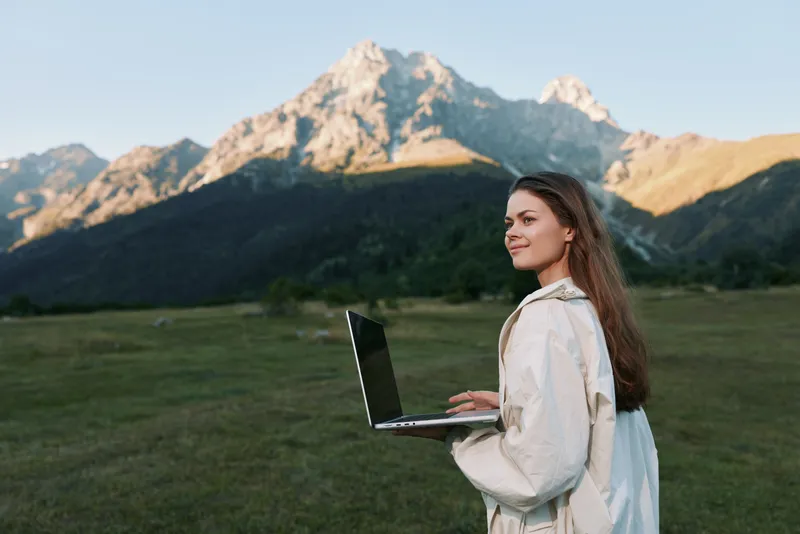 A cheerful woman stands in an open meadow with a laptop, wearing a light jacket, smiling and focused, enjoying outdoor work in a calm mountain landscape.