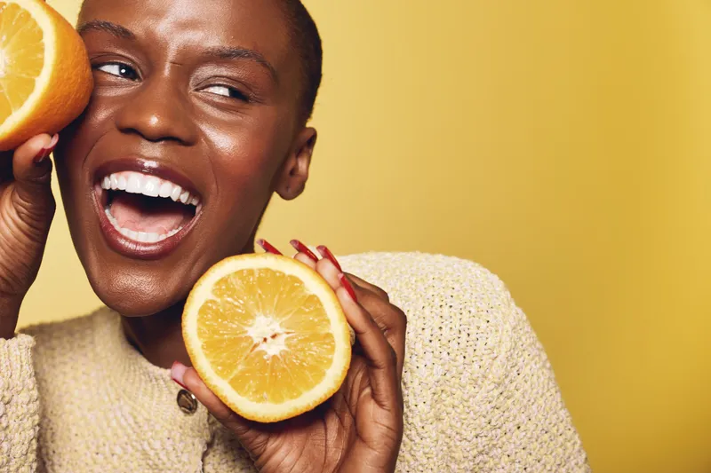 Happy dark skinned African woman with short hair holds two halves of fresh orange near face on yellow background. Concept of healthy lifestyle, fruit, nutrition, and natural beauty.