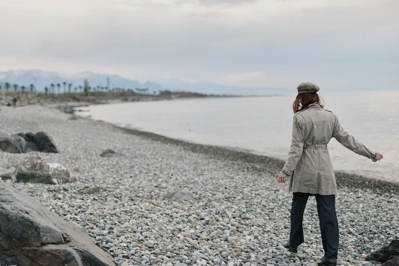 Woman in gray coat and hat walks on rocky beach near calm sea in cloudy weather. Concept of solitude, nature, and peaceful outdoor lifestyle during cool season.
