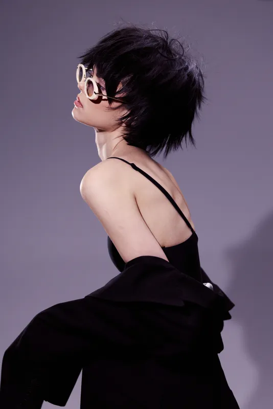 Stylish woman with short black hair wearing sunglasses and black clothing posing against purple background