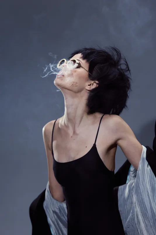 Smoking woman with glasses in black dress exhales smoke against gray background, stylish and moody atmosphere.