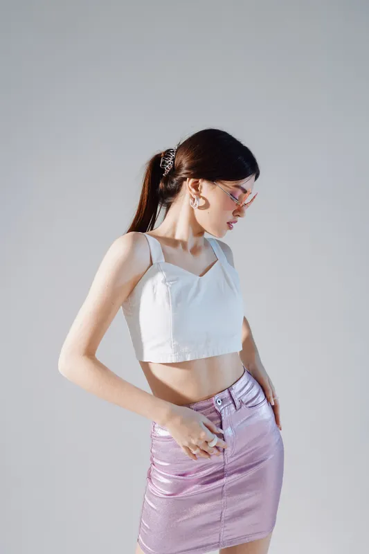 Young woman in a white crop top and shiny pink skirt poses sideways against a plain background. Concept of beauty, fashion, and modern style with minimalistic lighting.