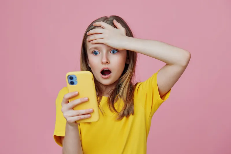 A shocked young girl in a bright yellow shirt reacts with disbelief while looking at her phone against a soft pink background, highlighting emotions of surprise and concern