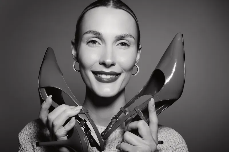 Smiling Caucasian woman with fair skin holding pair of high heel shoes close to face, studio portrait in black and white. Concept for fashion, style, and footwear.