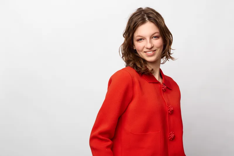 Smiling woman with short brown hair wearing a red knitted sweater stands against a white background. The casual portrait shows confidence, warmth, and relaxed mood in studio lighting.