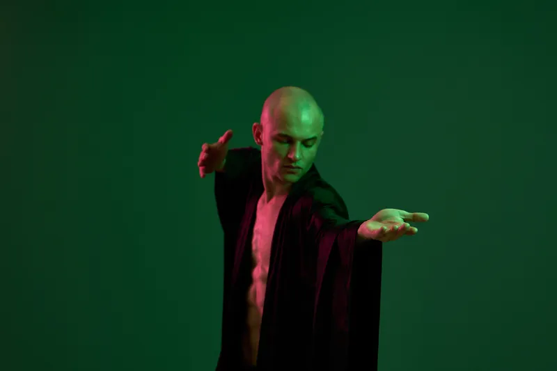 A man in a flowing black robe strikes a dynamic pose under soft green lighting. The composition highlights his movements and creates a striking visual contrast.