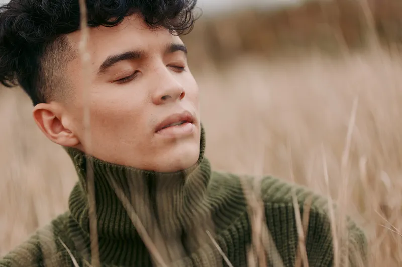 A serene portrait of a young person outdoors, eyes closed, skin lit warm, wearing a green knit turtleneck, conveying authentic presence and quiet credibility.