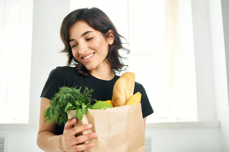 На кухне стоит жизнерадостная женщина с пакетом продуктов, что символизирует здоровое питание и домашние дела