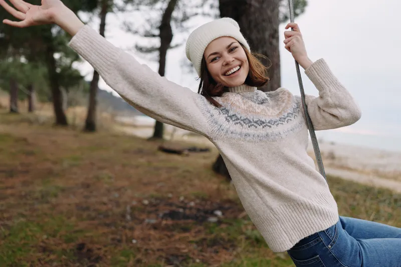 Woman on a swing wearing a cozy sweater and beanie, enjoying an outdoor park moment, smiling freely amid trees and open space, capturing casual autumn charm and playful mood