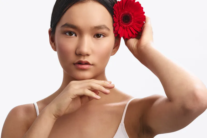 Asian woman with a red flower behind her ear gazes softly at the camera in a clean studio setting, conveying elegance and calm beauty.