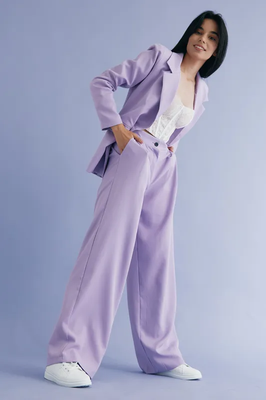 woman in lavender suit standing confidently against a plain purple background, wearing white sneakers and a lace top, fashion style portrait with soft lighting