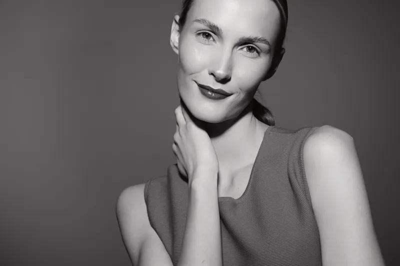 A Caucasian woman with fair skin and light hair poses wearing a sleeveless top in a soft monochrome studio setting. Concept for beauty, portrait, and fashion.