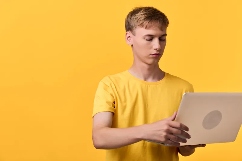 Man in a yellow t shirt holding a laptop against a yellow backdrop, showcasing modern technology, device usage, and focus on hands and screen