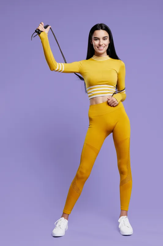 Young woman sport fitness wearing bright yellow activewear, holding a jump rope, smiling with confidence, standing against a purple background, healthy lifestyle concept.