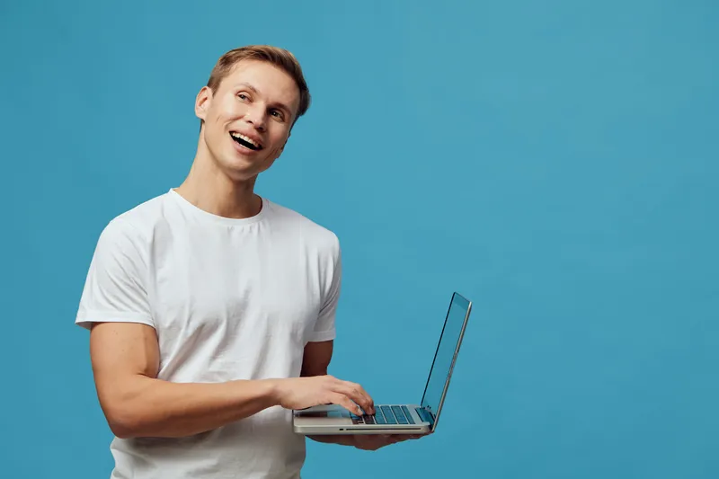 Smiling young man with short hair wearing casual white t-shirt holding open laptop computer isolated on solid blue background. Happy male with cheerful expression, technology use, workspace, positive 