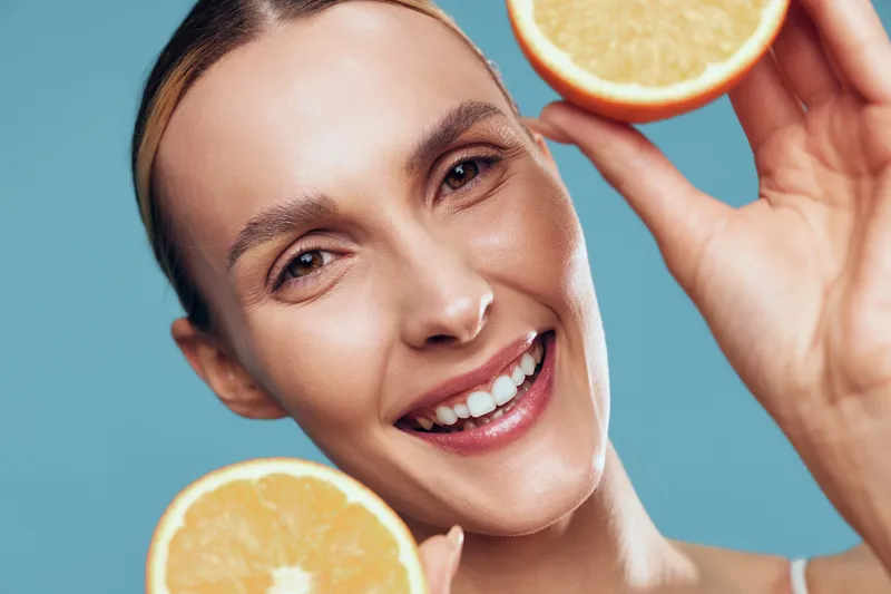 Smiling young woman with blonde hair holds two halves of a fresh orange near her face against a blue background expressing happiness and freshness.