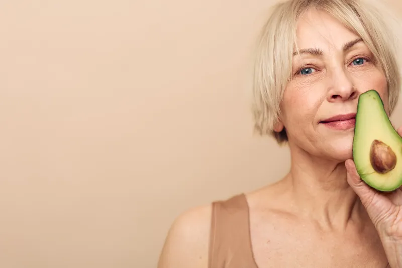 Smiling mature woman with short blonde hair holds fresh avocado half near face on beige background. Concept of healthy eating, natural skincare, wellness, and organic lifestyle for older adults.