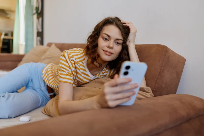 Relaxed woman lies on a sofa at home, looking at her smartphone, wearing a striped shirt, casual mood, cozy living room, soft light, candid lifestyle scene, peaceful moment