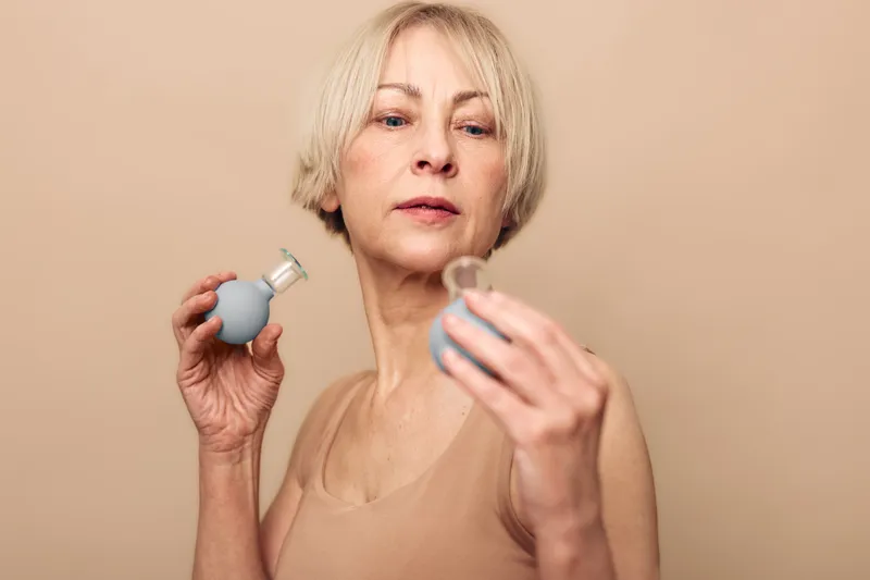 Mature woman with short blonde hair holding two glass cupping therapy devices, focusing on alternative wellness treatment in a neutral beige background studio setting.