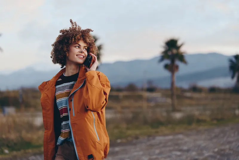 Woman lifestyle outdoors, smiling, orange jacket with denim, curly hair windblown, one hand touching hair, standing on rural road, mountain scenery with palm trees, natural sunlight