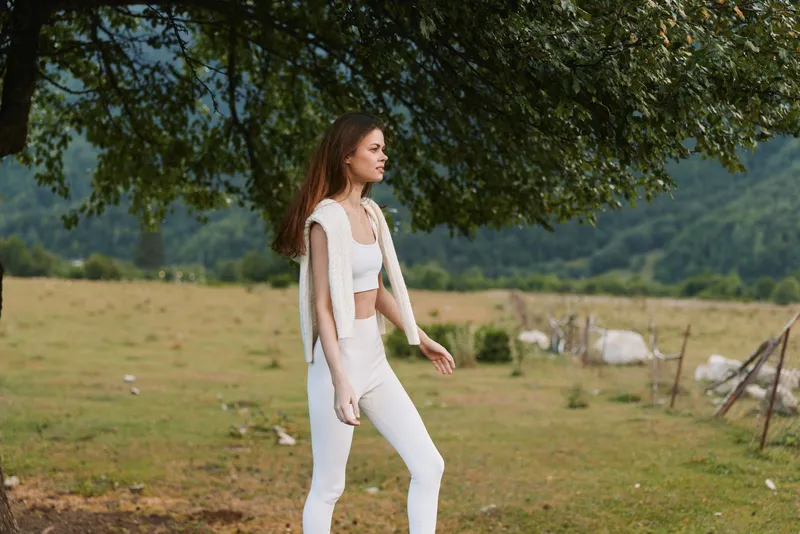 A woman in white casual outfit walks through a sunlit countryside field, capturing a relaxed lifestyle and serene mood amid open meadow, trees and hills, natural scenery.