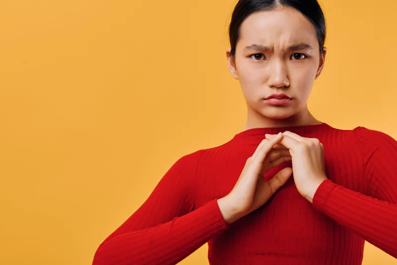 Young woman in red sweater showing heart shape with hands and serious facial expression isolated on yellow. Portrait of girl making gesture for love and care concept with focus.