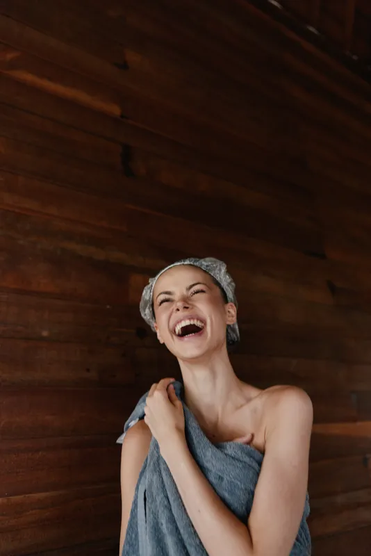 laughing woman wrapped in towel with shower cap enjoys relaxing moment indoors against warm wooden wall background expressing joy and freshness