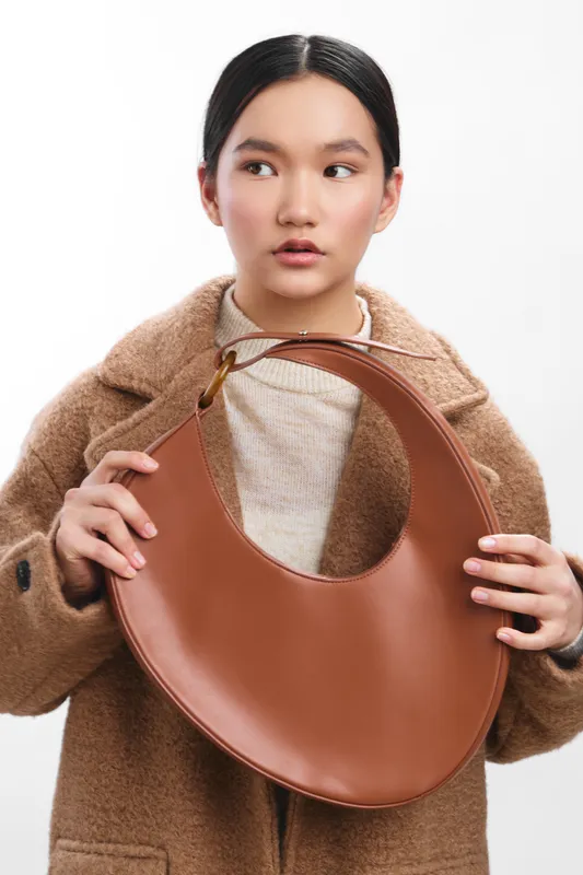 asian woman with a calm expression holding a large brown circular handbag, wearing a beige knit top and tan coat, posing in a studio against a white backdrop.