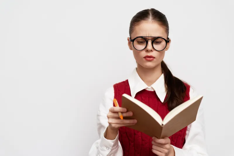 Молодая женщина в очках сосредоточенно читает коричневую книгу, держа в руке оранжевую ручку