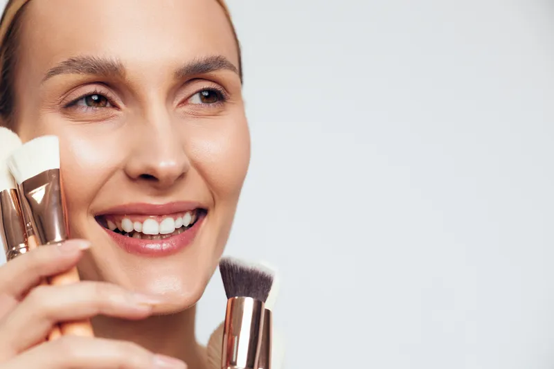 Smiling woman holding makeup brushes close to her face on a white background. Concept of beauty, cosmetics, skincare, and professional makeup application with soft natural lighting.