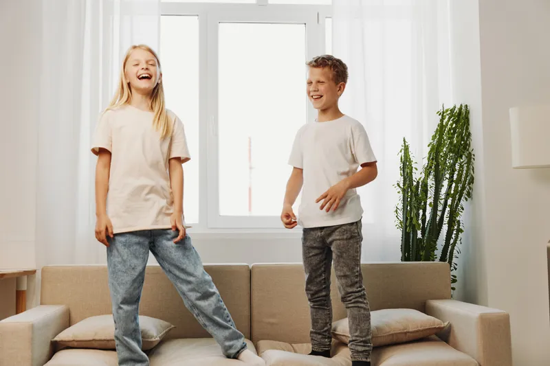 Happy children jumping on sofa in bright living room, laughing and enjoying playful moment together at home with natural light.