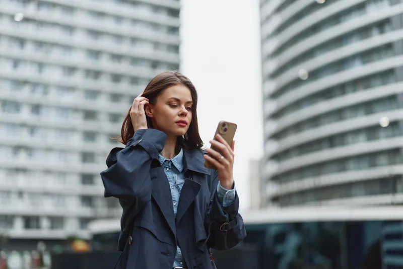 Young woman in dark coat uses smartphone outdoors in modern city environment with curved glass buildings, focused on screen, casual urban lifestyle concept.