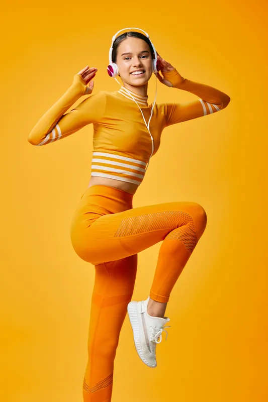 Energetic woman in bright orange athletic wear jumping with headphones against a vivid orange backdrop, showcasing fitness, dance and active lifestyle in a bold studio setting.