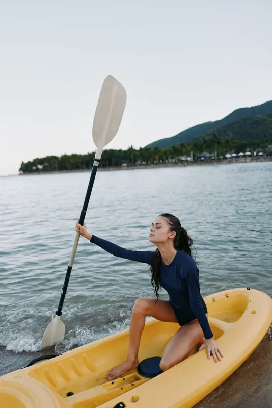Young woman in a blue swimsuit sits in a yellow kayak on calm water, lifting a paddle as waves roll toward a pebble shore and distant hills frame the scene.