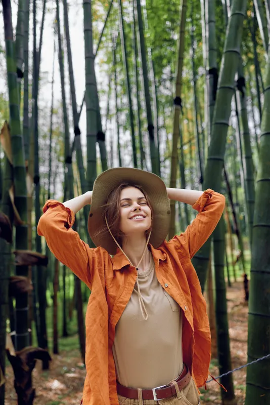 Woman with hat and smile in a bamboo forest, nature portrait of relaxed outdoor traveler wearing orange shirt and boho outfit, enjoying peaceful green environment and sunlight.