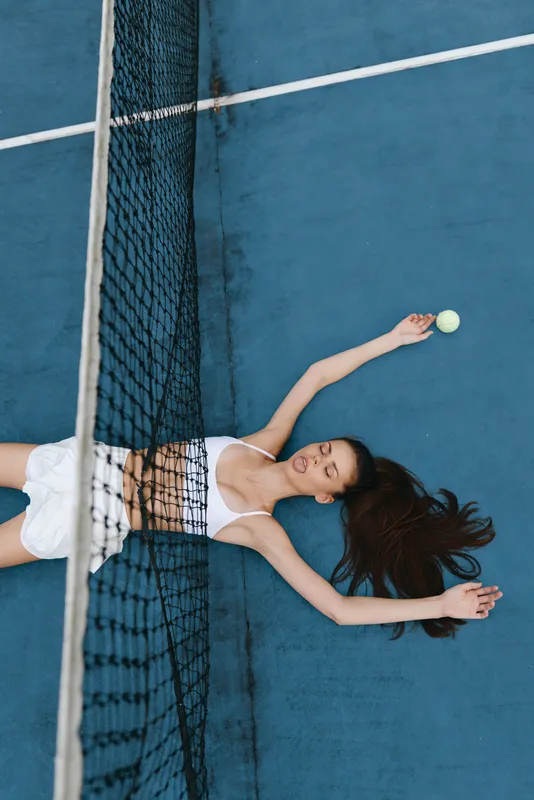 Young woman celebrating victory on a tennis court, wearing sporty attire and showing excitement with arms raised, with a blue background and tennis net