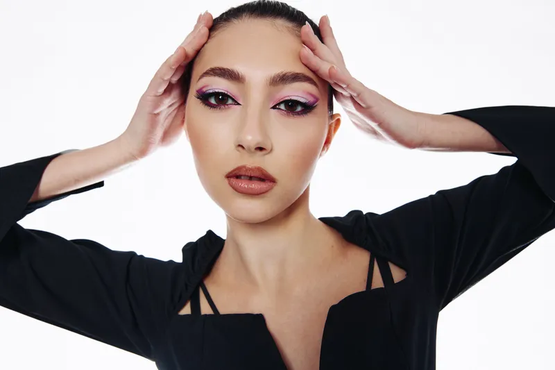 Fashion model with creative pink eye makeup and nude lips poses confidently against a white background, highlighting bold beauty and contemporary style in a studio portrait.