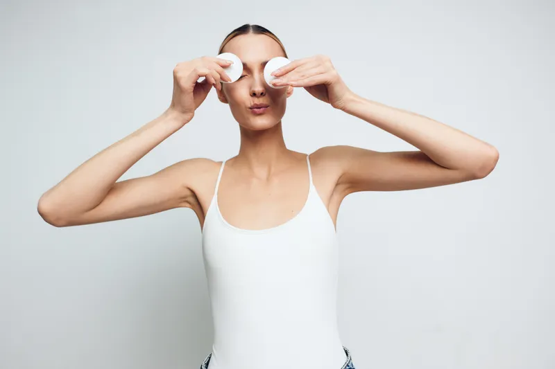 Young woman in white tank top holding cotton pads over eyes with playful expression on light background. Concept of skincare, beauty, and self-care routine with natural lighting.