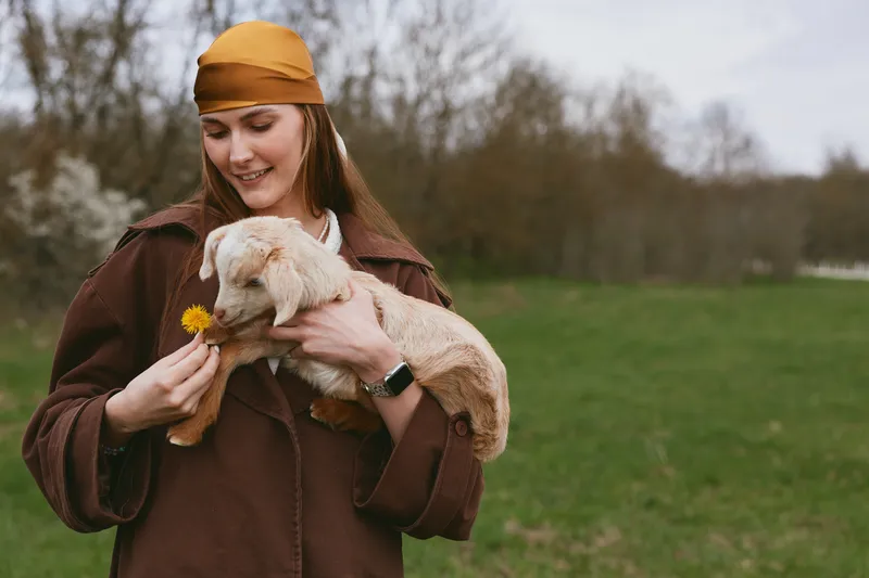 Joyful woman outdoors pets a gentle goat kid, holds a dandelion, sharing a warm moment of affection in a pastoral field, rustic jacket and scarf, serene countryside backdrop, candid lifestyle scene.