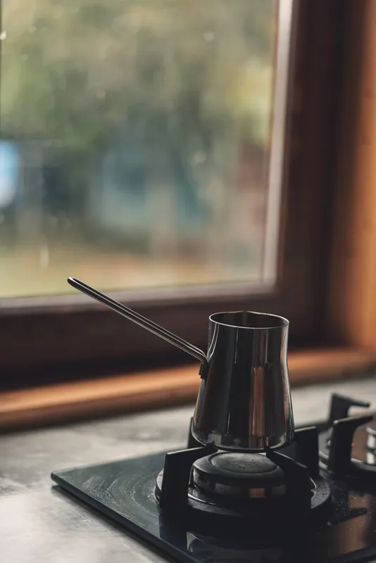 Moka pot brews on a compact stove beside a sunlit window, metal shine and steam shaping a cozy morning ritual in a simple kitchen