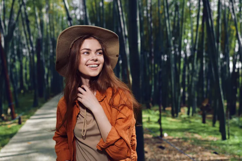 Woman smile outdoors wearing hat and orange jacket on a forest path, walking and enjoying nature, portrait of a young adult traveler with candid happy expression and warm sunlight.