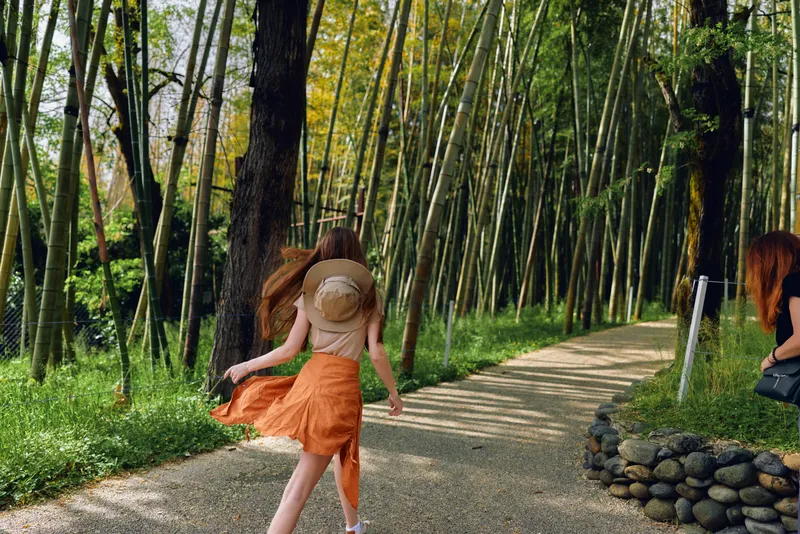 Woman girl running on a gravel path through a bamboo forest wearing a straw hat and orange skirt, nature travel scene with sunlight, carefree walking and exploration in green grove