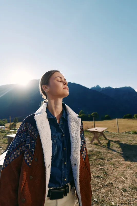 Woman outdoors in mountains with sunlight on her face, portrait of nature and jacket-clad figure standing peaceful with eyes closed, serene rural lifestyle moment in a meadow.