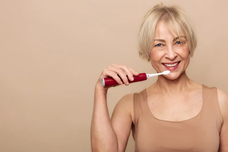 Smiling mature woman in beige tank top brushing teeth with electric toothbrush, looking at camera. Concept of dental hygiene, oral care, healthy smile, and personal routine.