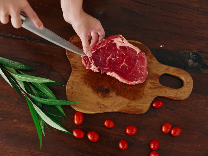Женщина с ножом в руке режет свежий стейк из мяса для жарки на кухне