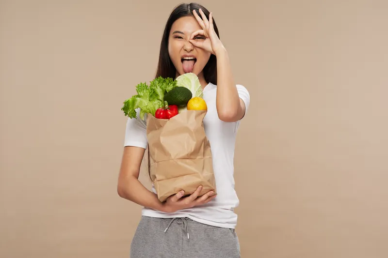 Женщина с пакетом продуктов держит два пальца возле лица на бежевом фоне