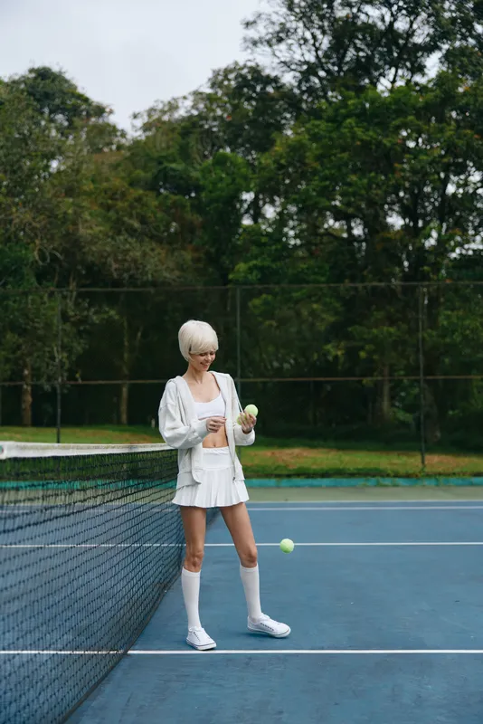 Stylish female tennis player in athletic wear, smiling while holding tennis balls on a court surrounded by greenery, embodying the spirit of outdoor sports