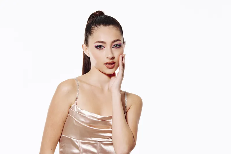 Young woman in rose gold satin dress poses touching face with elegant makeup and pulled back hair. Bright studio light on white background. Fashion and beauty concept.