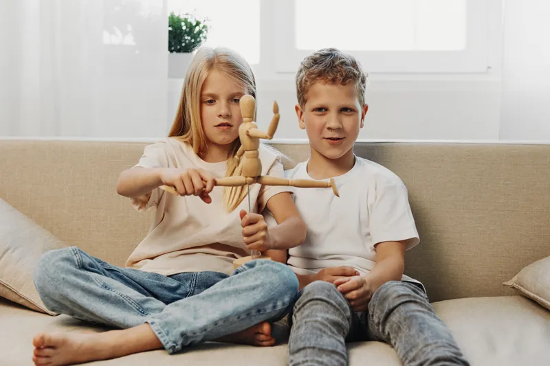 Two children sitting on a sofa holding a wooden artist mannequin together in a bright living room. Concept of creativity, childhood, play, and education at home.