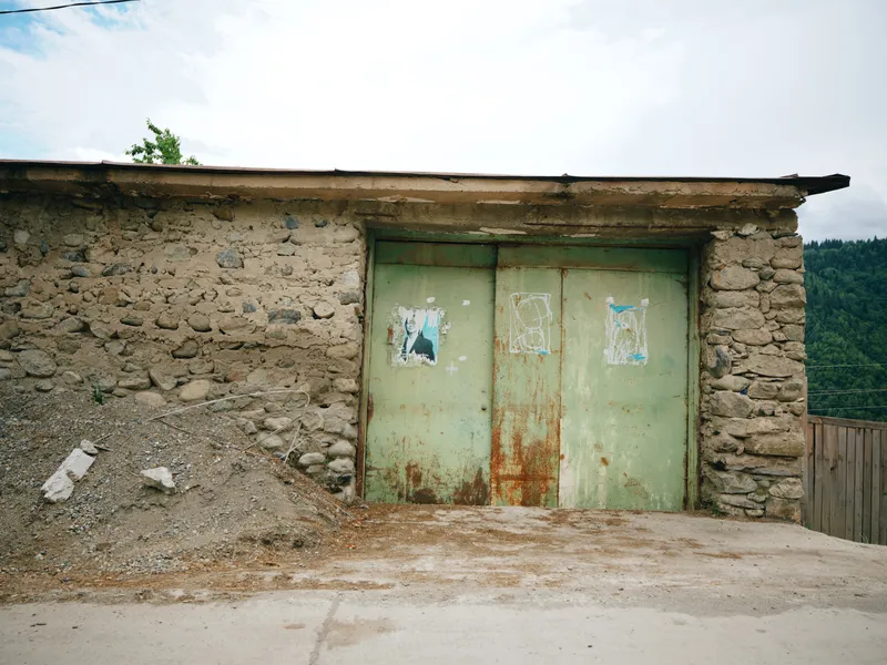 Rustic shed facade with weathered green doors, stone walls, dirt ground and hillside backdrop, abandoned rural building with earthy tones, textured exterior and simple construction